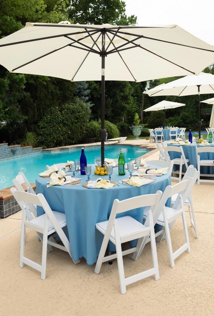 Round tables in light blue linens under white market umbrellas beside a pool